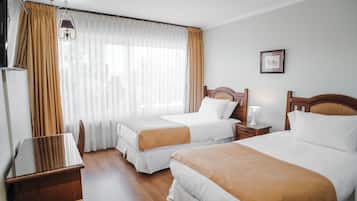 Habitación de hotel con dos camas, un escritorio de madera, una silla, una mesita de noche, una lámpara y un cuadro en la pared.