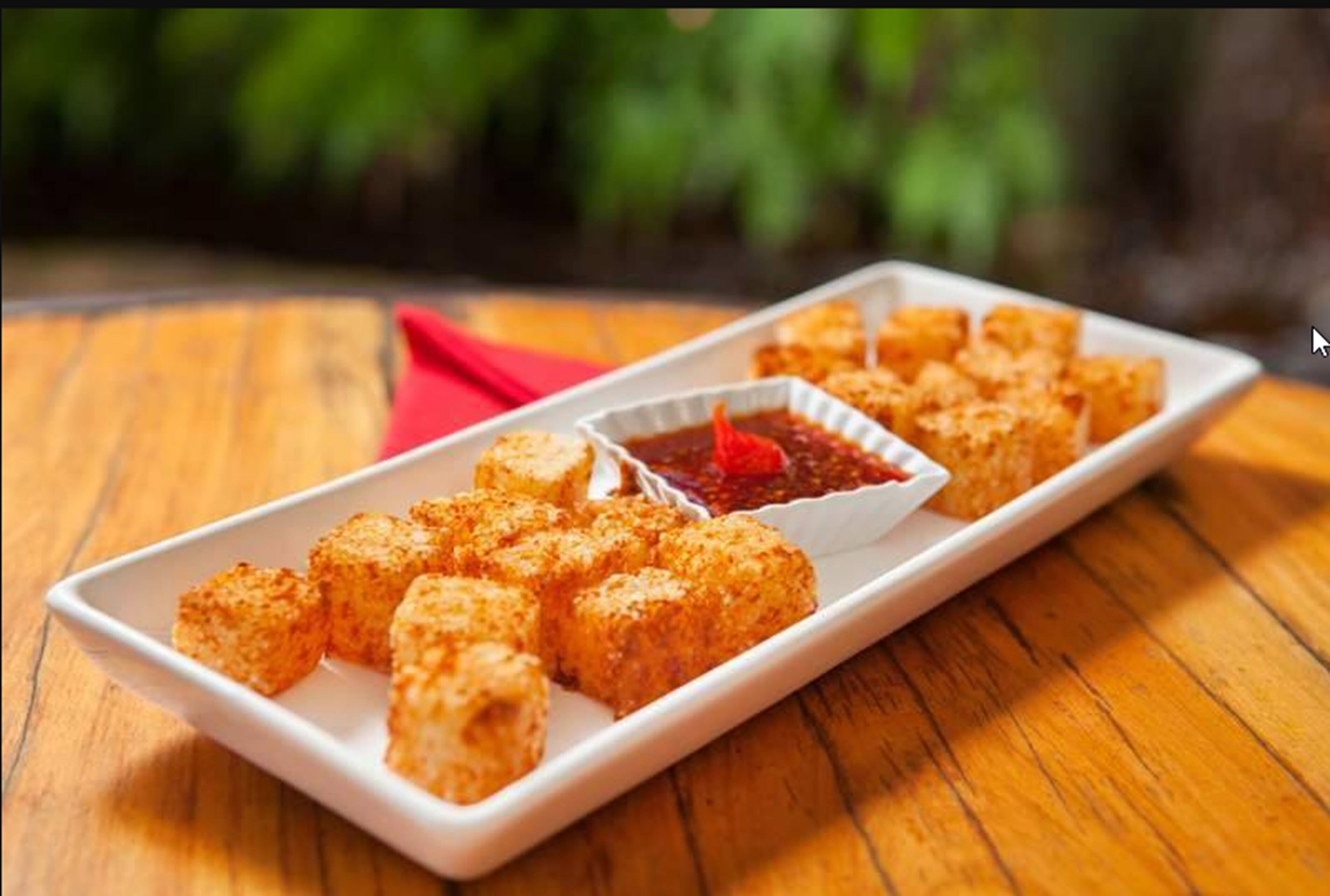 A rectangular white plate with cubed food items and a small bowl of red sauce on a wooden table.