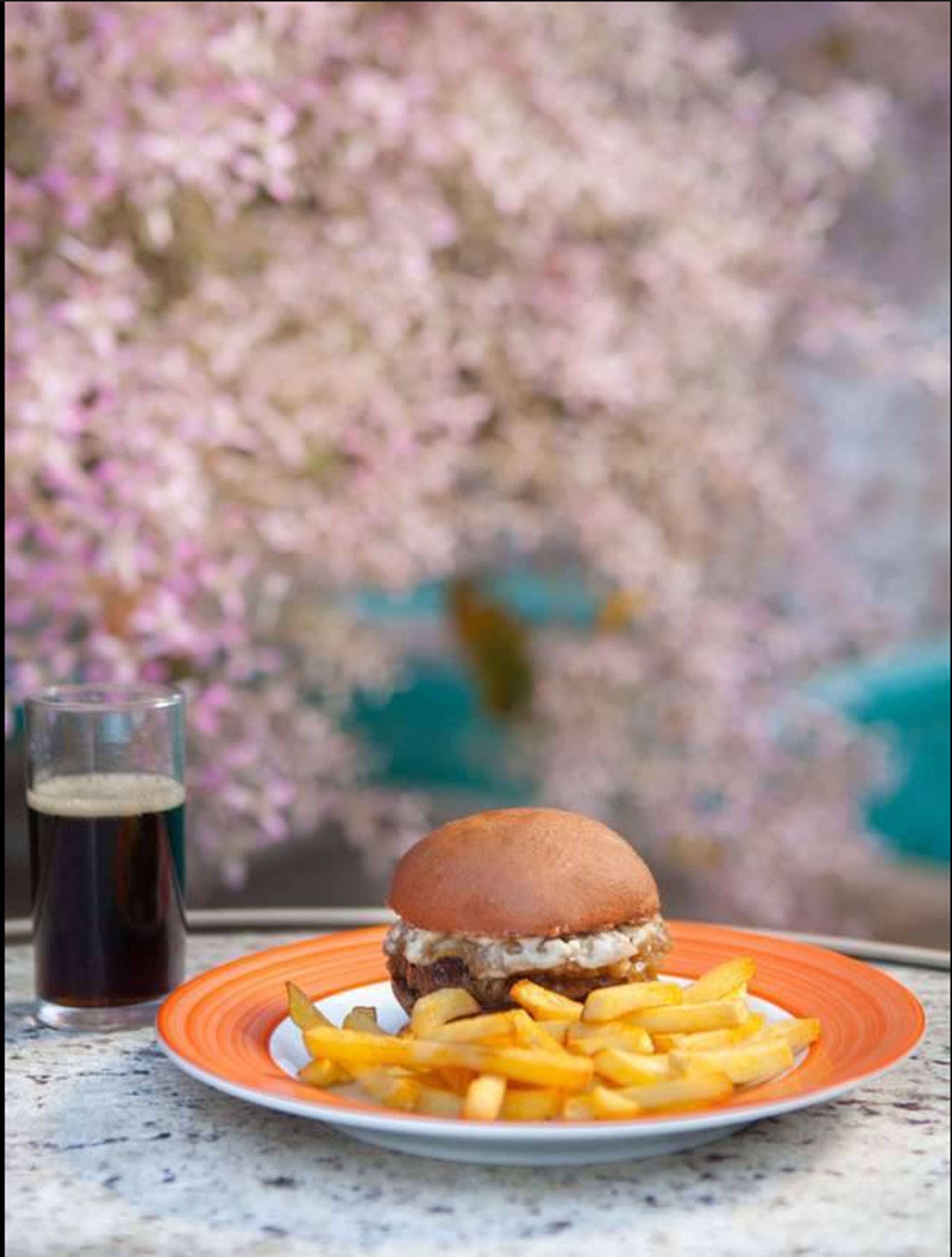 A burger with fries on a plate, a glass of dark beverage, and a blurred background of pink flowers.