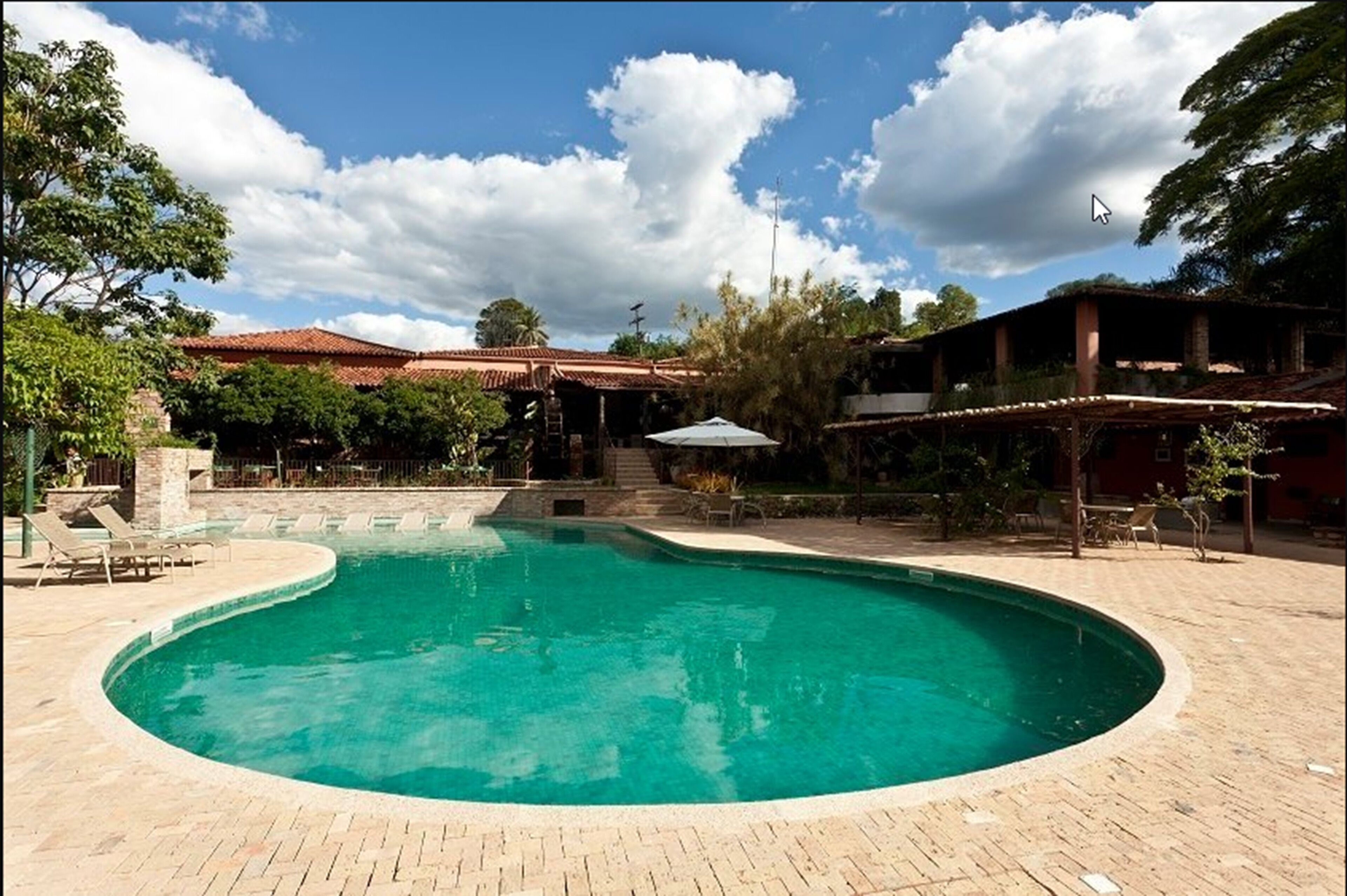 A swimming pool with a curved edge, surrounded by a brick patio. There are lounge chairs and a covered seating area.