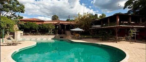 A swimming pool with a curved edge, surrounded by a brick patio. There are lounge chairs and a covered seating area.