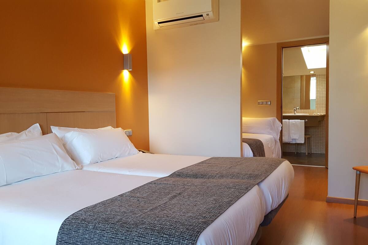 Quarto de hotel com duas camas, banheiro com espelho e uma cadeira.