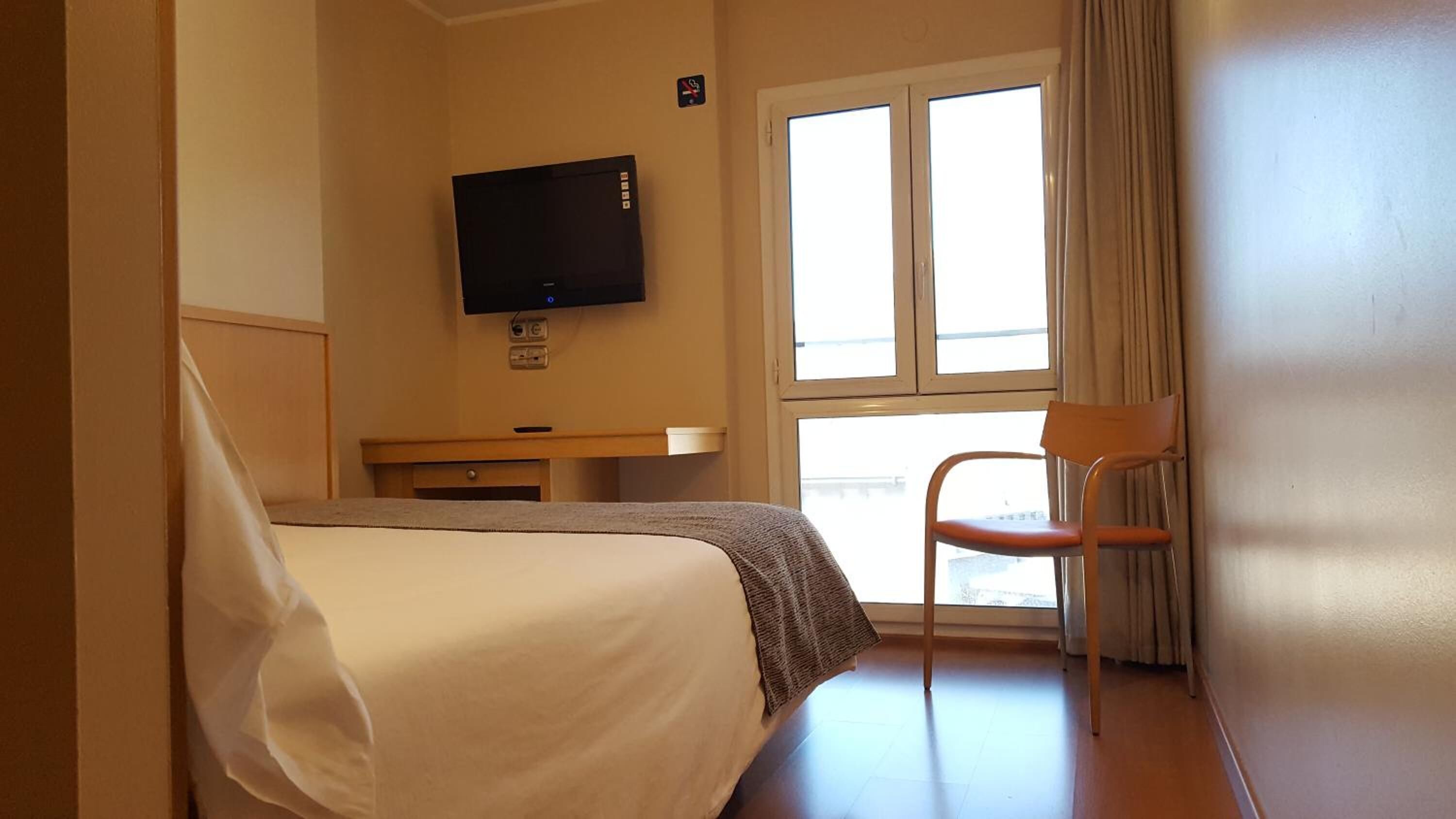 A hotel room with a bed, a television mounted on the wall, a window with curtains, and a chair.