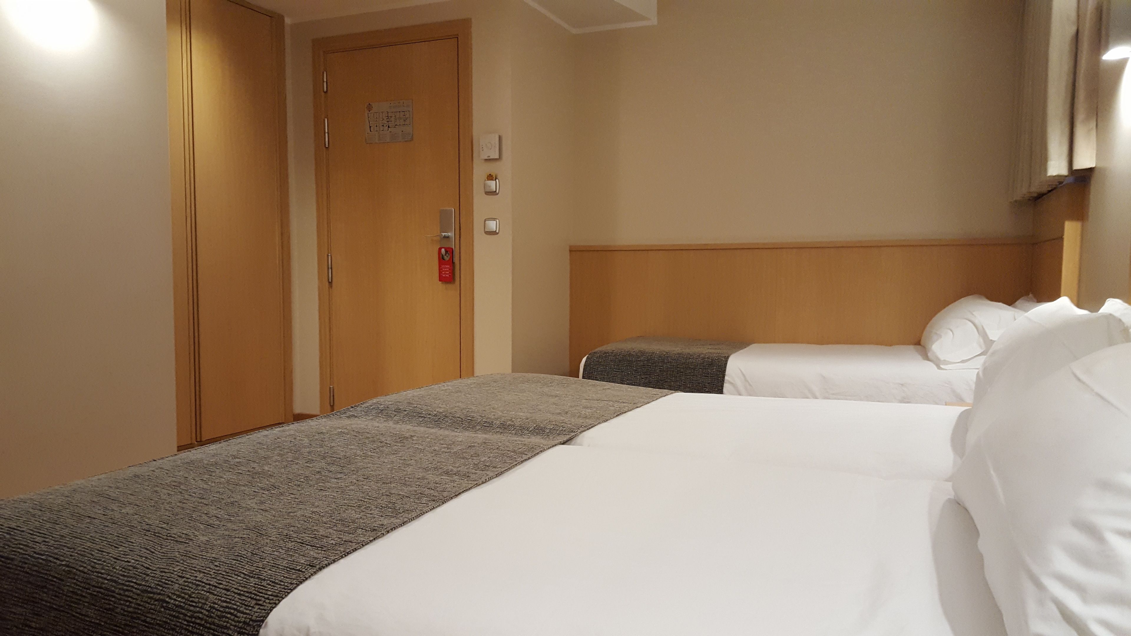 A hotel room with two beds, a wooden wardrobe, a door with a fire alarm, and a thermostat on the wall.