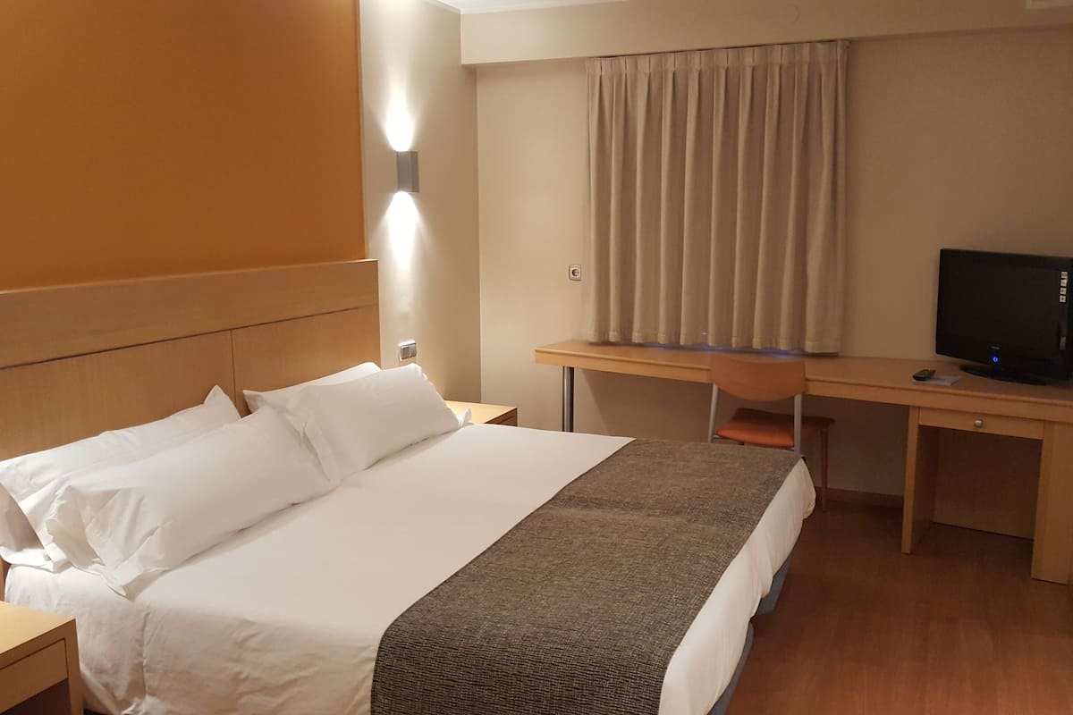 Quarto de hotel com uma cama, escrivaninha, cadeira e televisão.