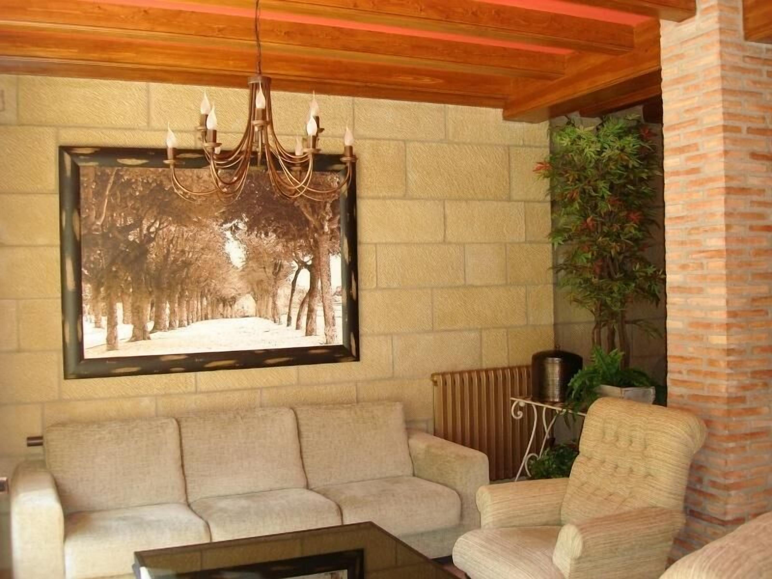A living room with a beige sofa, a patterned armchair, a coffee table, a framed picture of trees, a hanging chandelier, and a brick wall.
