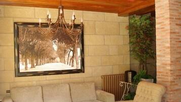 A living room with a beige sofa, a patterned armchair, a coffee table, a framed picture of trees, a hanging chandelier, and a brick wall.