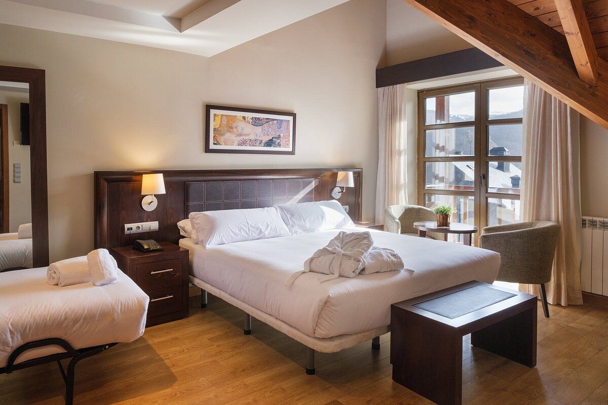 A hotel room with a large bed, a bedside table with a lamp, a chair, and a view of the mountains through a window.