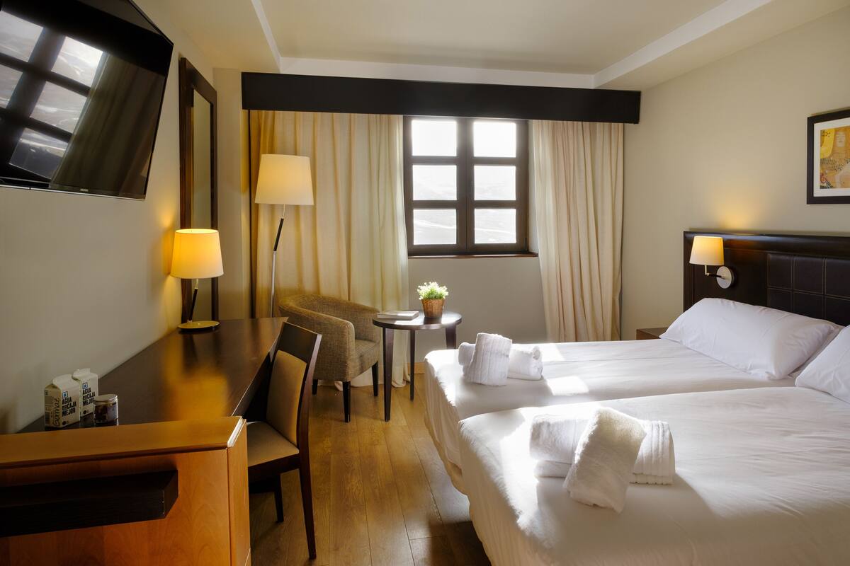 A hotel room with two beds, a desk, a chair, a TV, and a window with curtains.