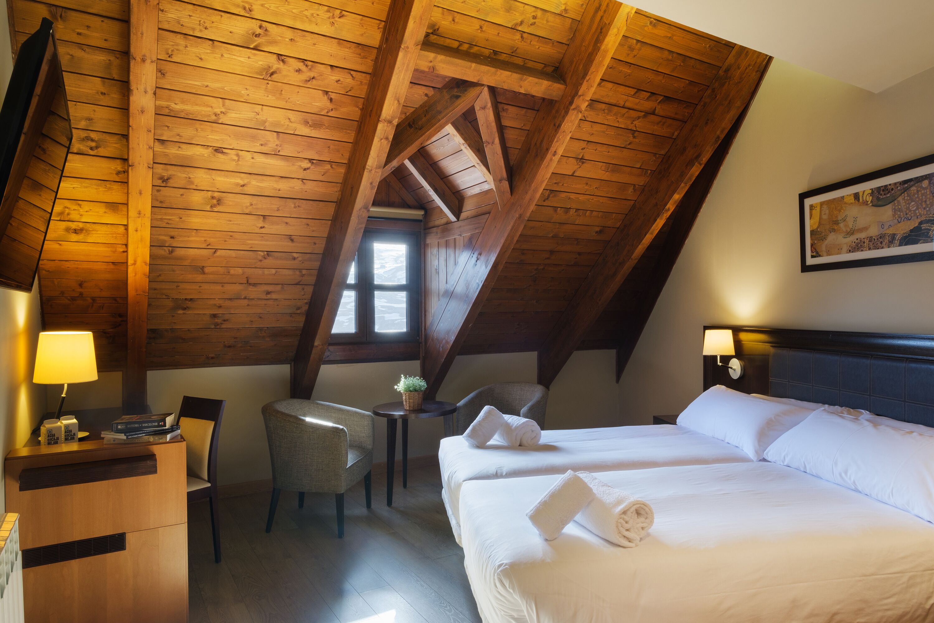 Una habitación con techo inclinado, vigas de madera, una cama con ropa blanca, un escritorio con lámpara, una silla y una pequeña mesa con una planta.