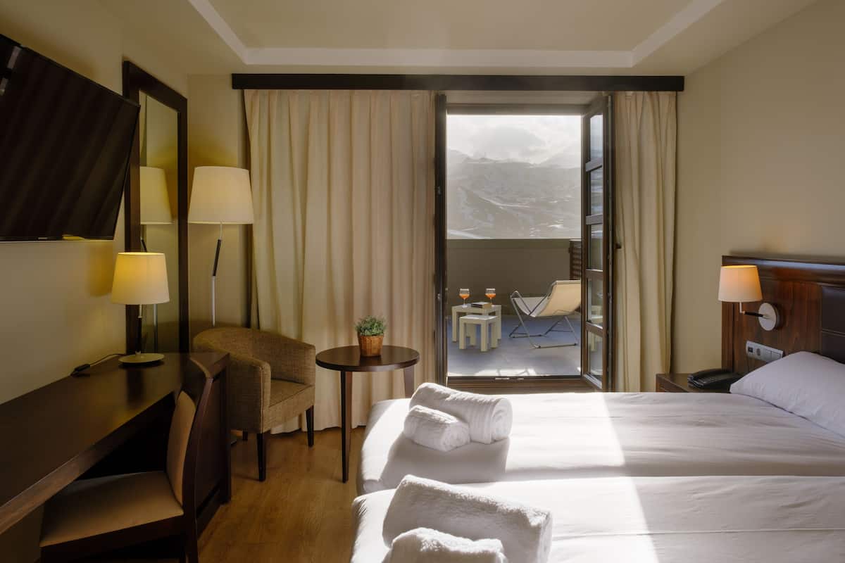 A hotel room with a bed, a desk, a chair, a small table with a plant, and a view of the mountains through the open door.