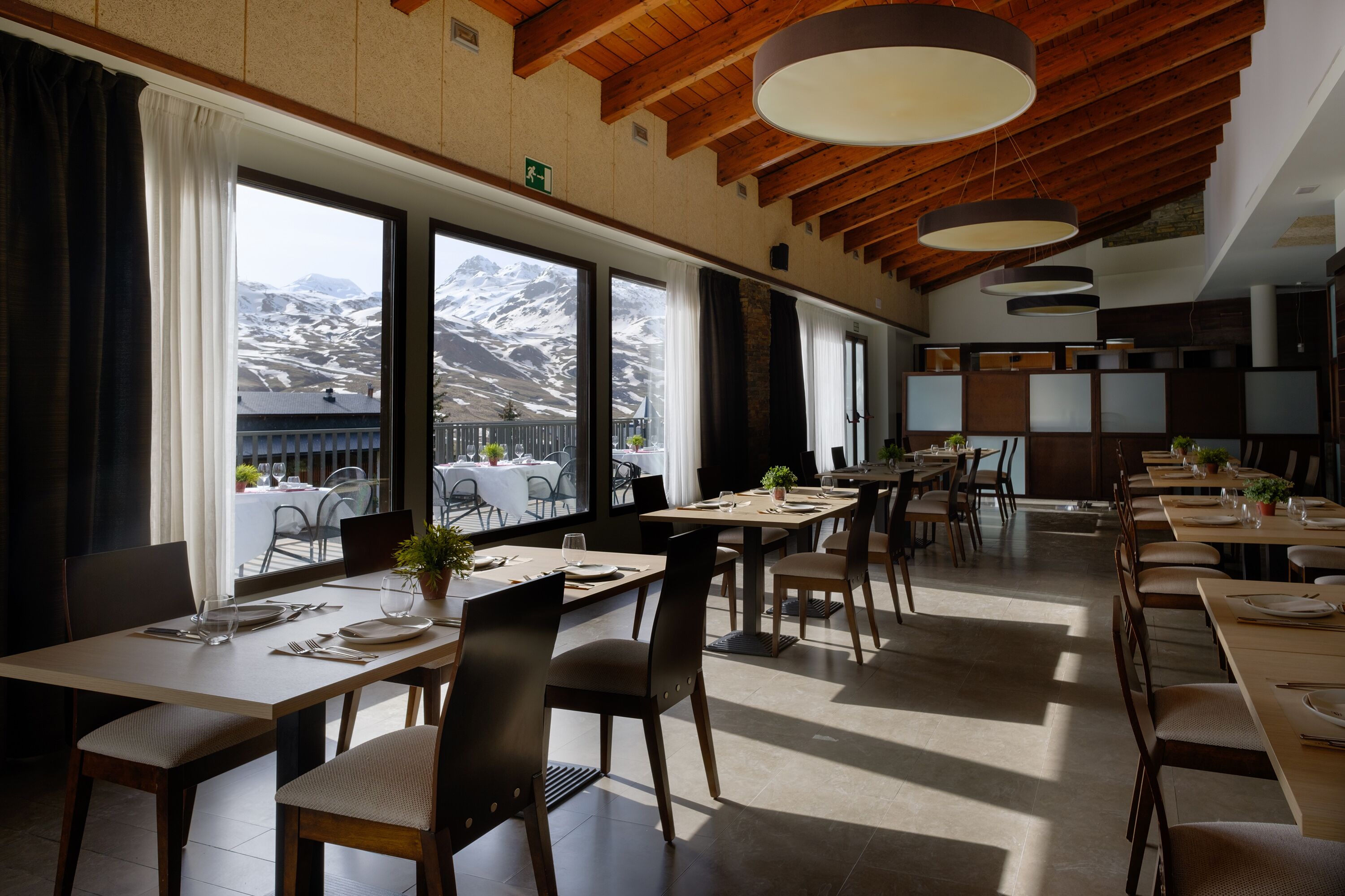 A restaurant with multiple tables and chairs, large windows with a mountain view, and wooden ceiling beams.