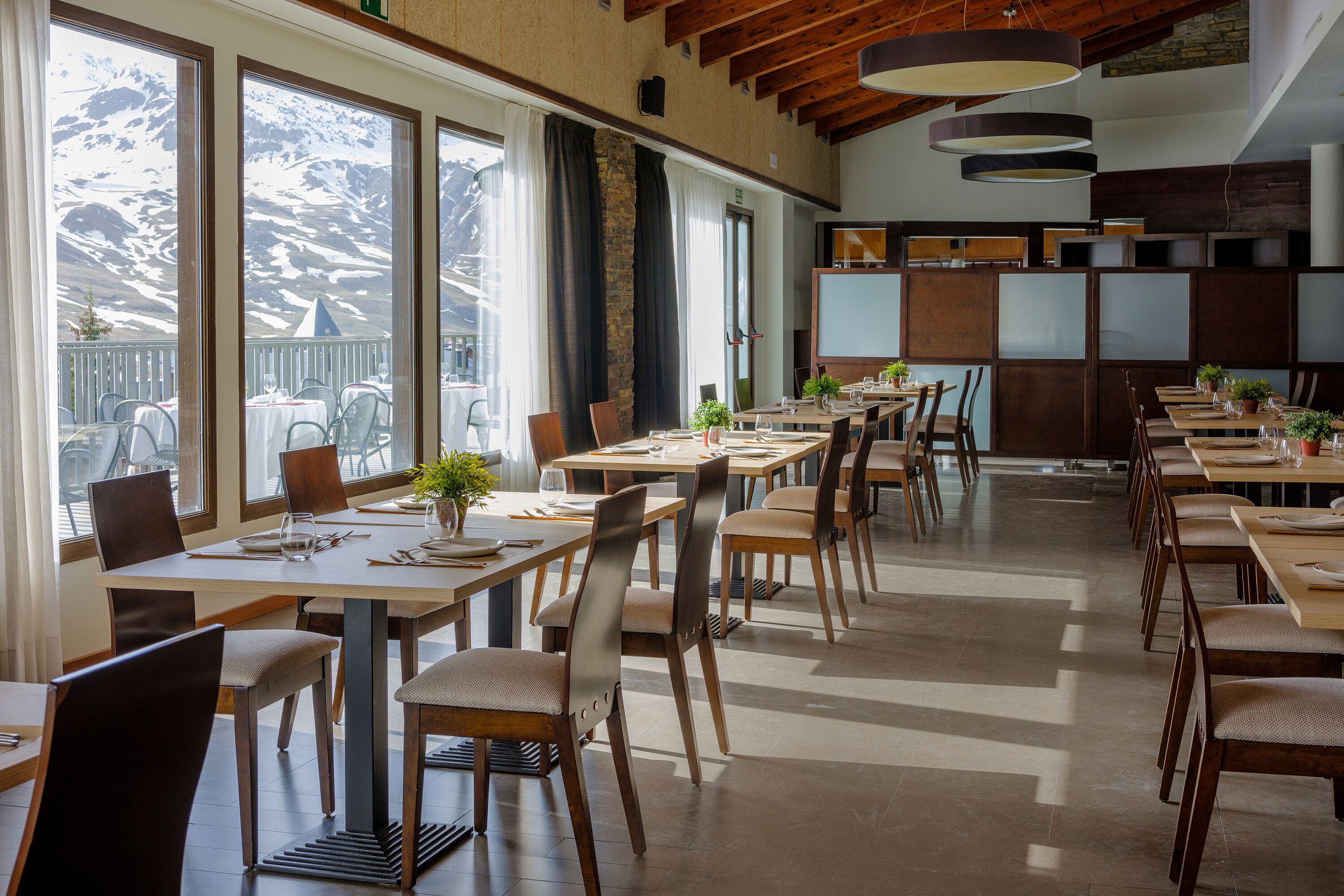 A restaurant with multiple tables and chairs, large windows with a mountain view, and wooden ceiling beams.