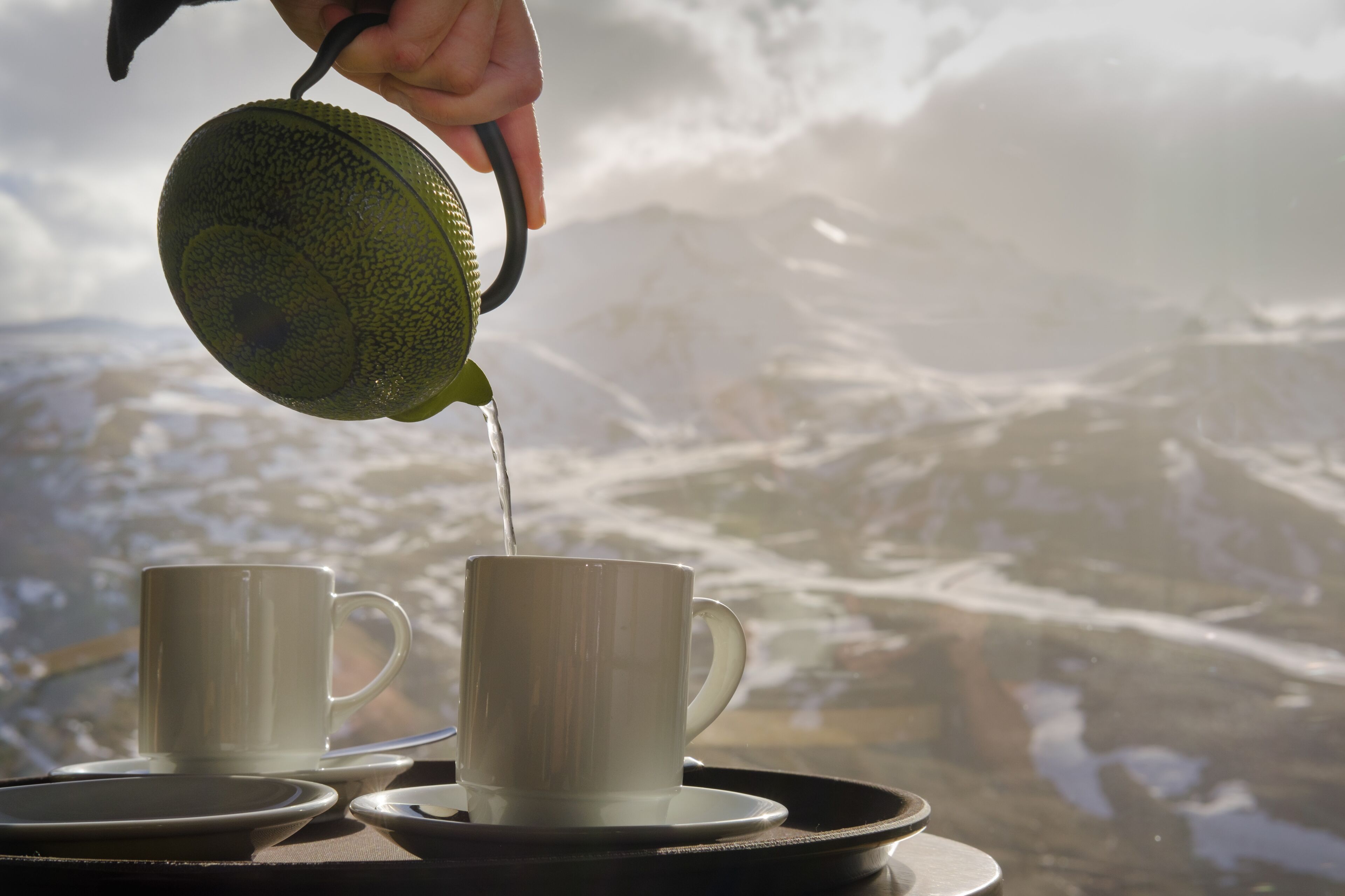 A hand pouring tea into a white cup, with a scenic mountain view in the background.