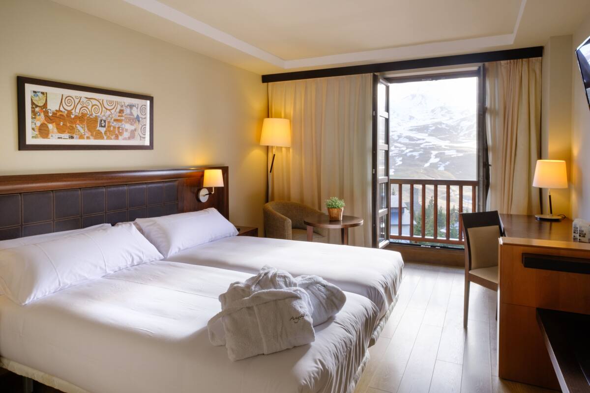 A hotel room with a large bed, two bedside tables, a desk, a chair, a small table with a plant, and a window with curtains.