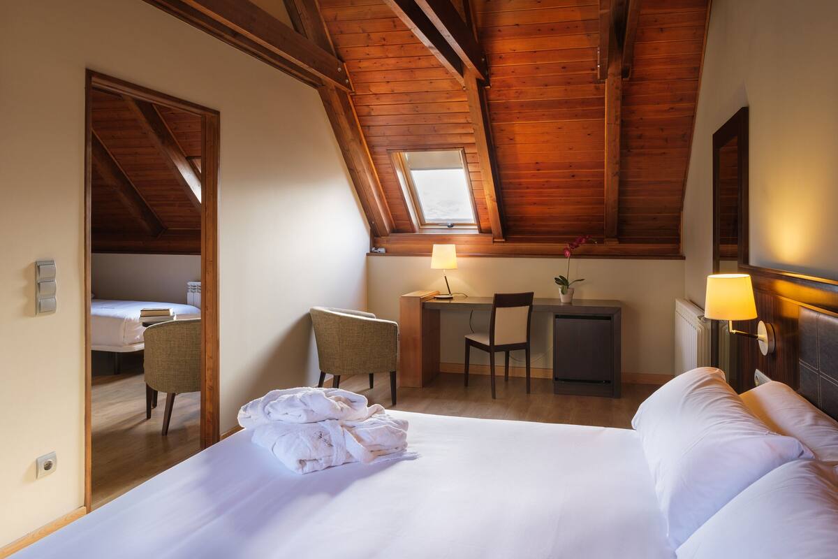 A hotel room with a large bed, a desk, a chair, and a small table with a lamp.