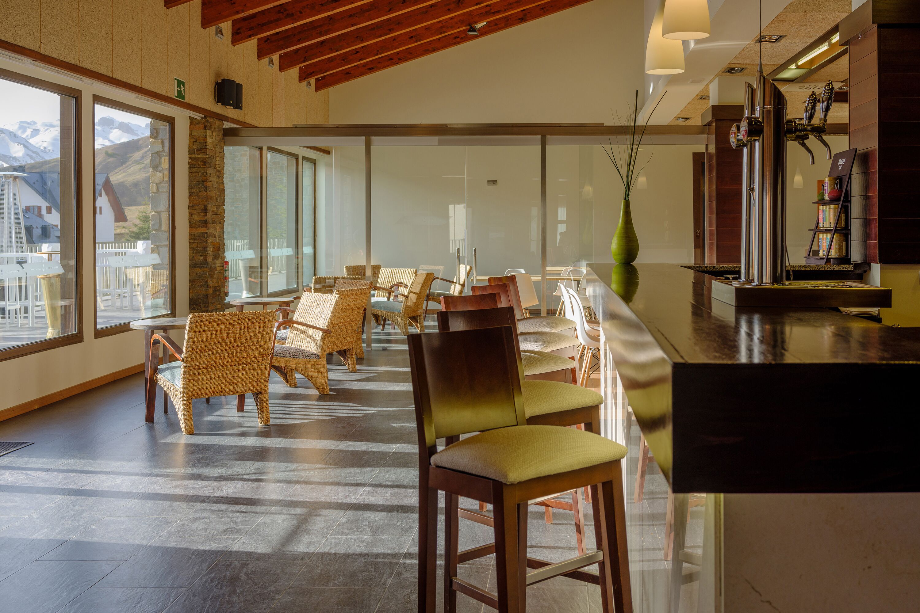 Un interior modern amb cadires de vimet, una zona de bar i grans finestres que ofereixen vista a la muntanya.