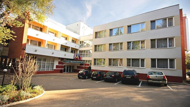 Ein Hotel mit zentralem Eingang, mehreren Stockwerken und einem Parkplatz mit verschiedenen Autos.