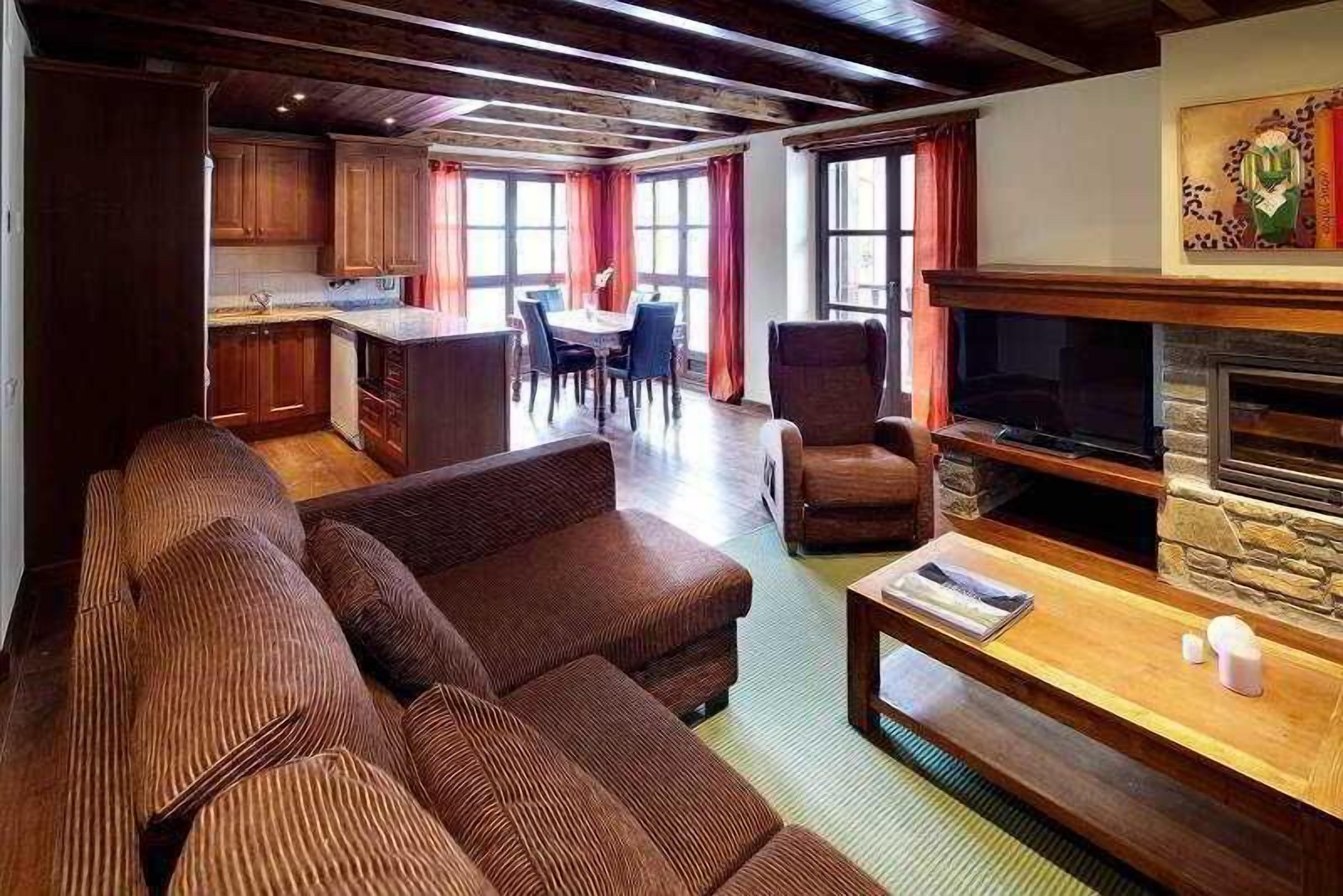 A spacious living area with a large sofa, a television, and a fireplace. There is a dining table and chairs in the background, and a kitchen area is visible to the left.