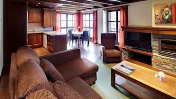 A spacious living area with a large sofa, a television, and a fireplace. There is a dining table and chairs in the background, and a kitchen area is visible to the left.