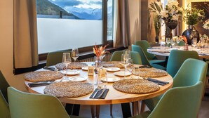 Comedor con mesas redondas dispuestas para una comida, amplias ventanas con vista a la montaña y sillas tapizadas en tono verde.