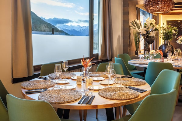 A dining area with round tables set for a meal, large windows with a mountain view, and green upholstered chairs. - Meliá de Tredós (Naut Aran)