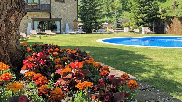 Un edificio de piedra de dos pisos con piscina, rodeado por un césped cuidado y flores coloridas.