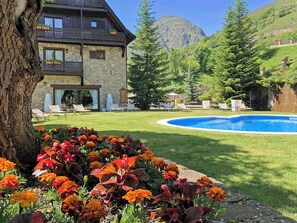 Un edificio de piedra de dos pisos con piscina, rodeado por un césped cuidado y flores coloridas.