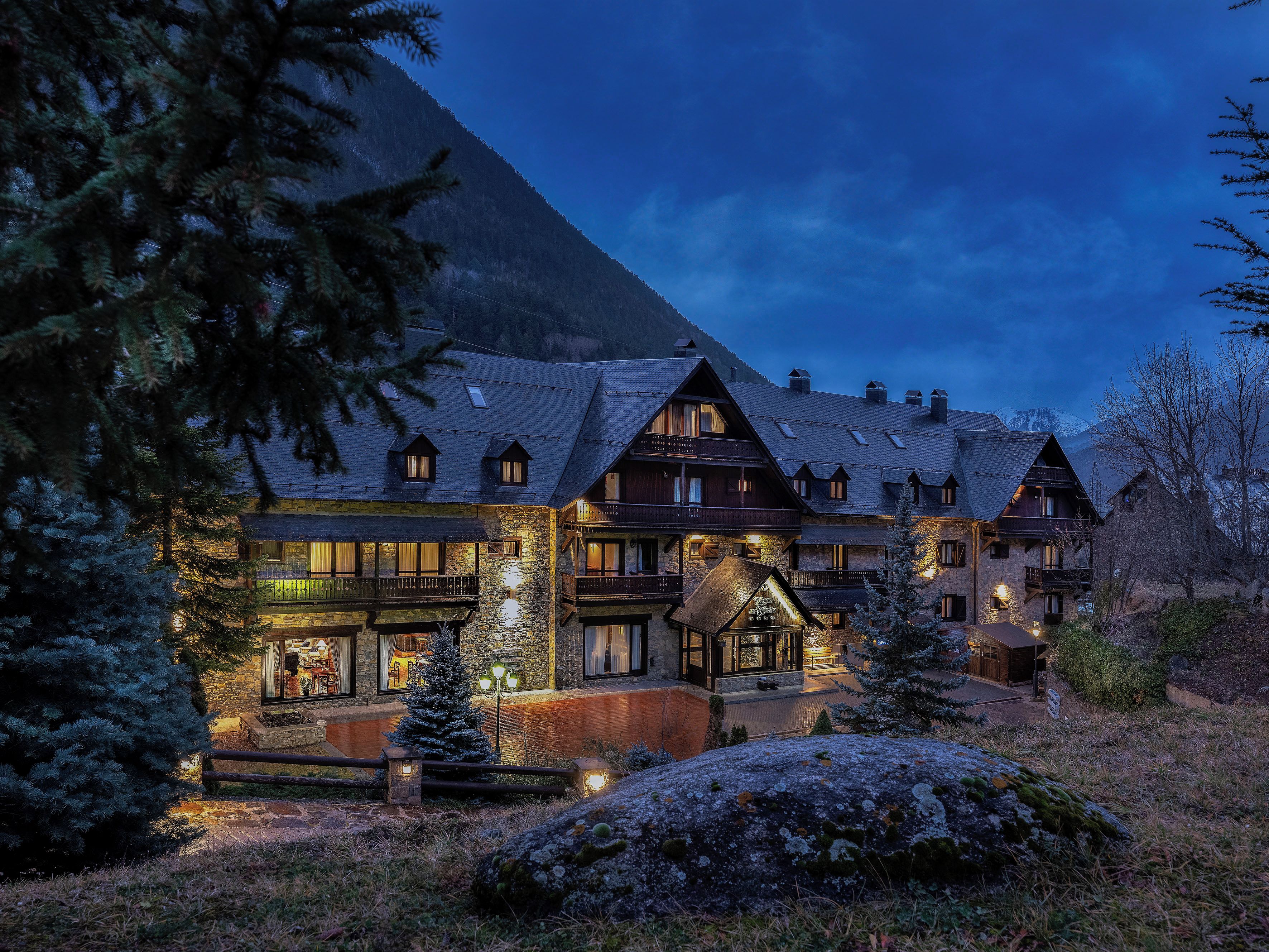 A traditional chalet-style hotel with multiple balconies and a stone facade, surrounded by snow-covered trees and a mountainous backdrop.