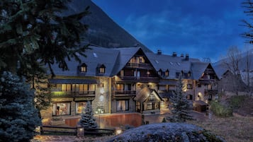 A traditional chalet-style hotel with multiple balconies and a stone facade, surrounded by snow-covered trees and a mountainous backdrop.