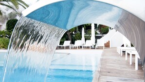 A pool area with a waterfall feature, wooden deck, and white lounge chairs. - Apartamentos Castaví (Formentera)