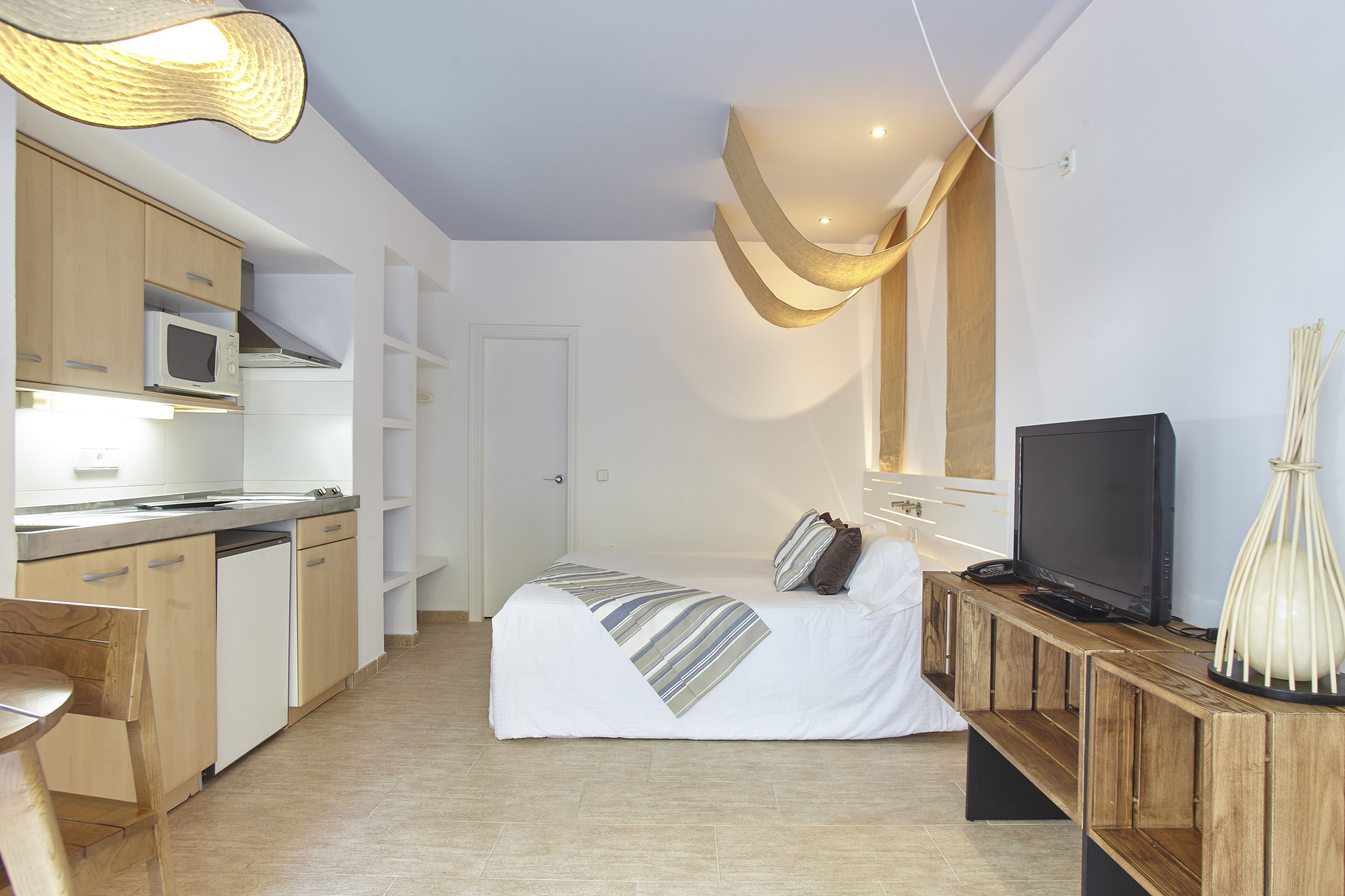 A compact hotel room with a kitchenette, a bed with a striped blanket, a television, and a unique ceiling light fixture.