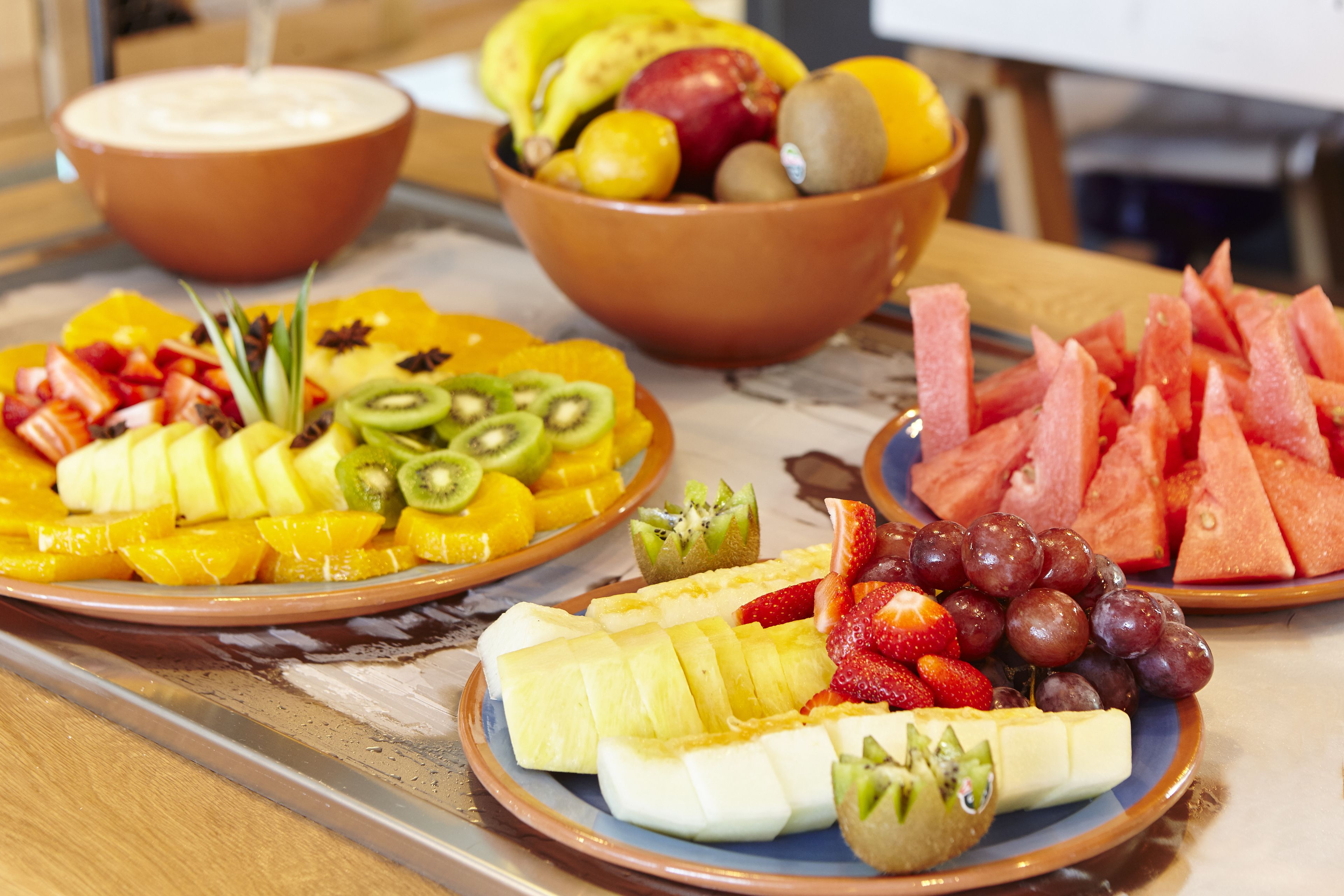 Una mesa con diferentes bandejas de fruta, incluyendo plátanos en rodajas, manzanas, naranjas y uvas.