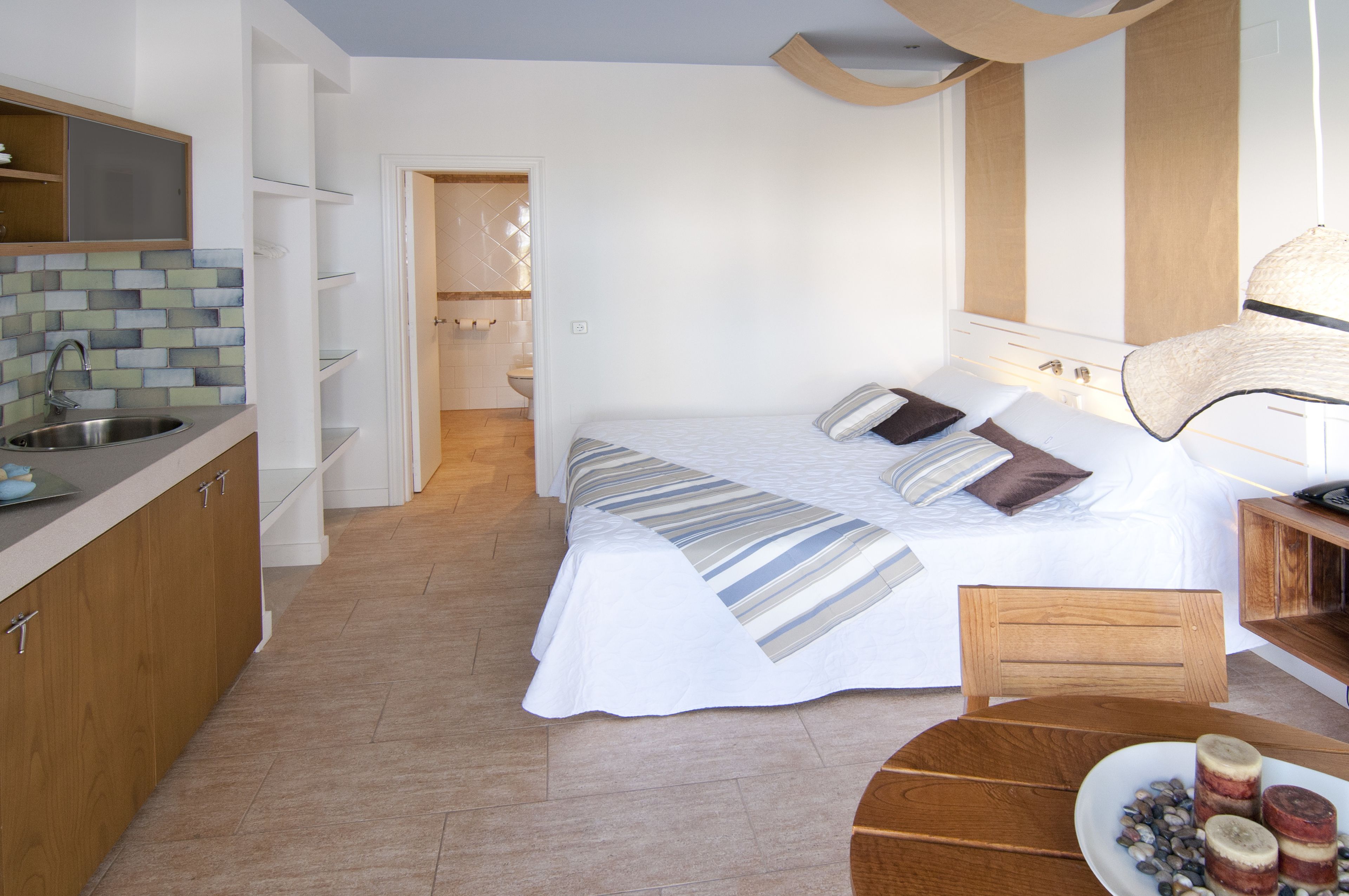 A compact hotel room with a bed, bedside table, and a small round table with a plate of food. A kitchen area with a sink and cabinets is visible in the background.