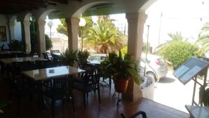 An indoor dining area with tables and chairs, potted plants, and a view of the street outside. - Oasis D'or (Santanyi)