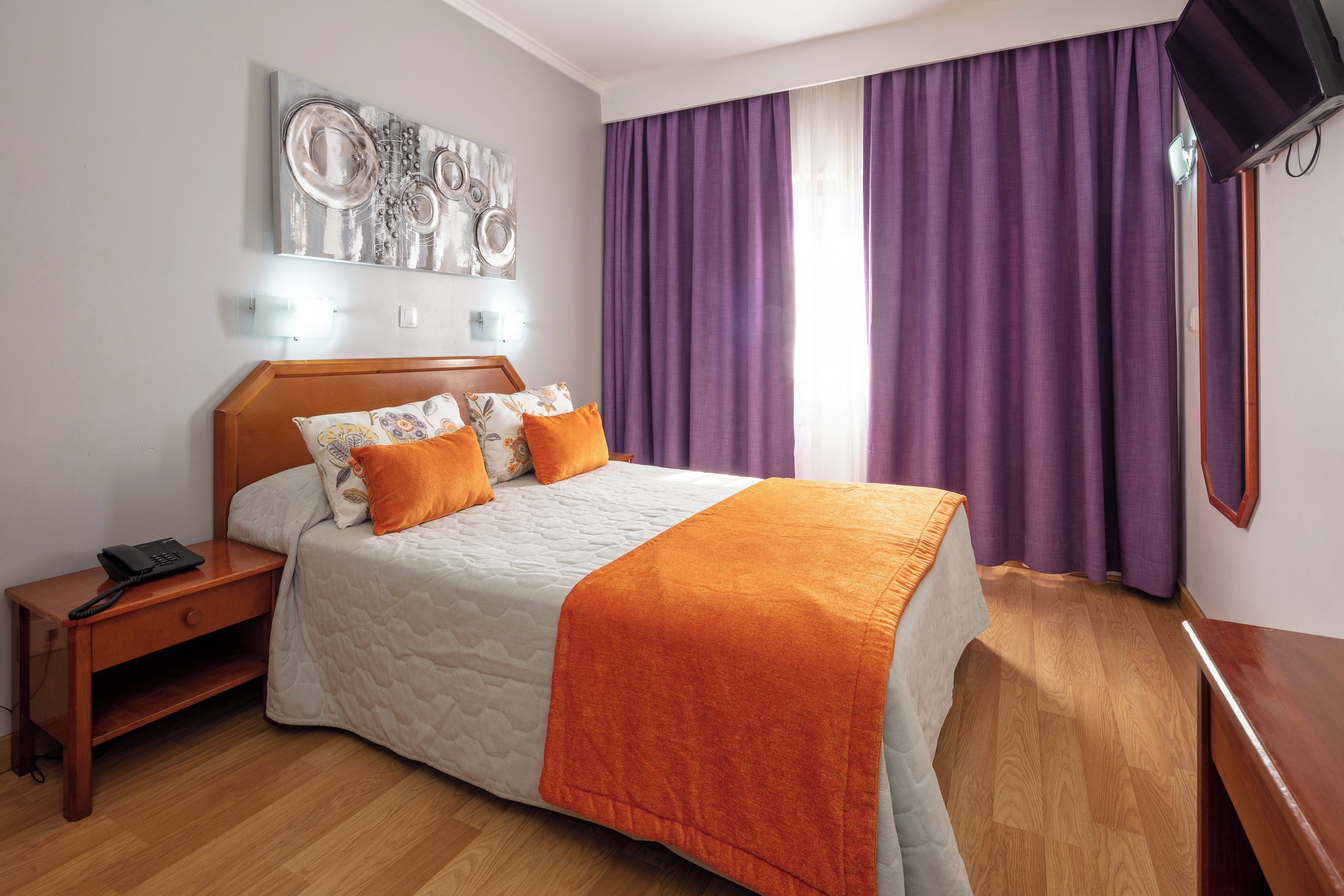 A neatly arranged bedroom with a bed, a bedside table, a wall-mounted artwork, and purple curtains.