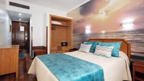 Um quarto com cama, escrivaninha, guarda-roupa e um mural com um pôr do sol sobre o oceano.