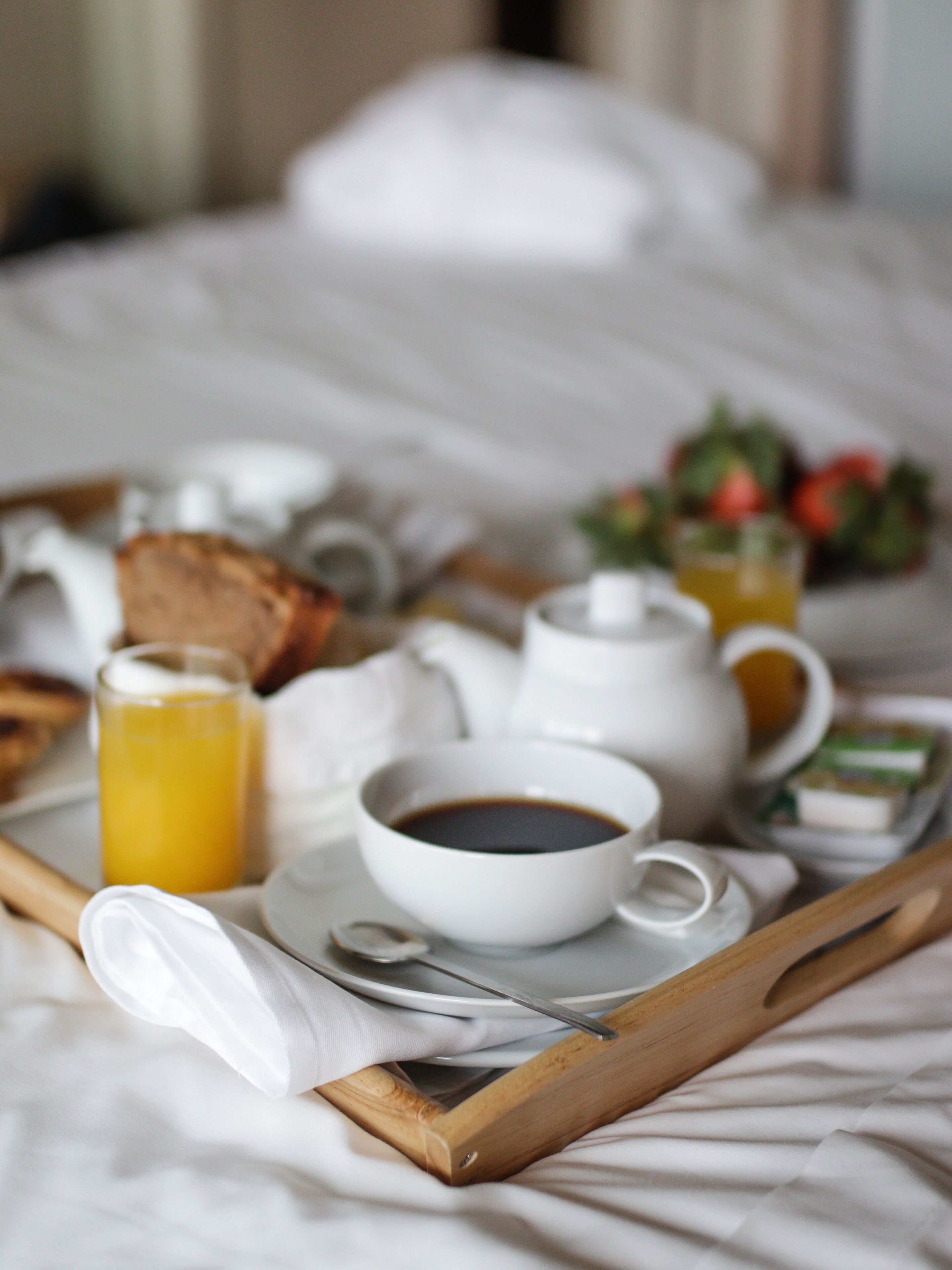 A breakfast tray with a cup of coffee, a glass of orange juice, a teapot, a teacup, a plate with toast, and a small bowl of strawberries on a bed.