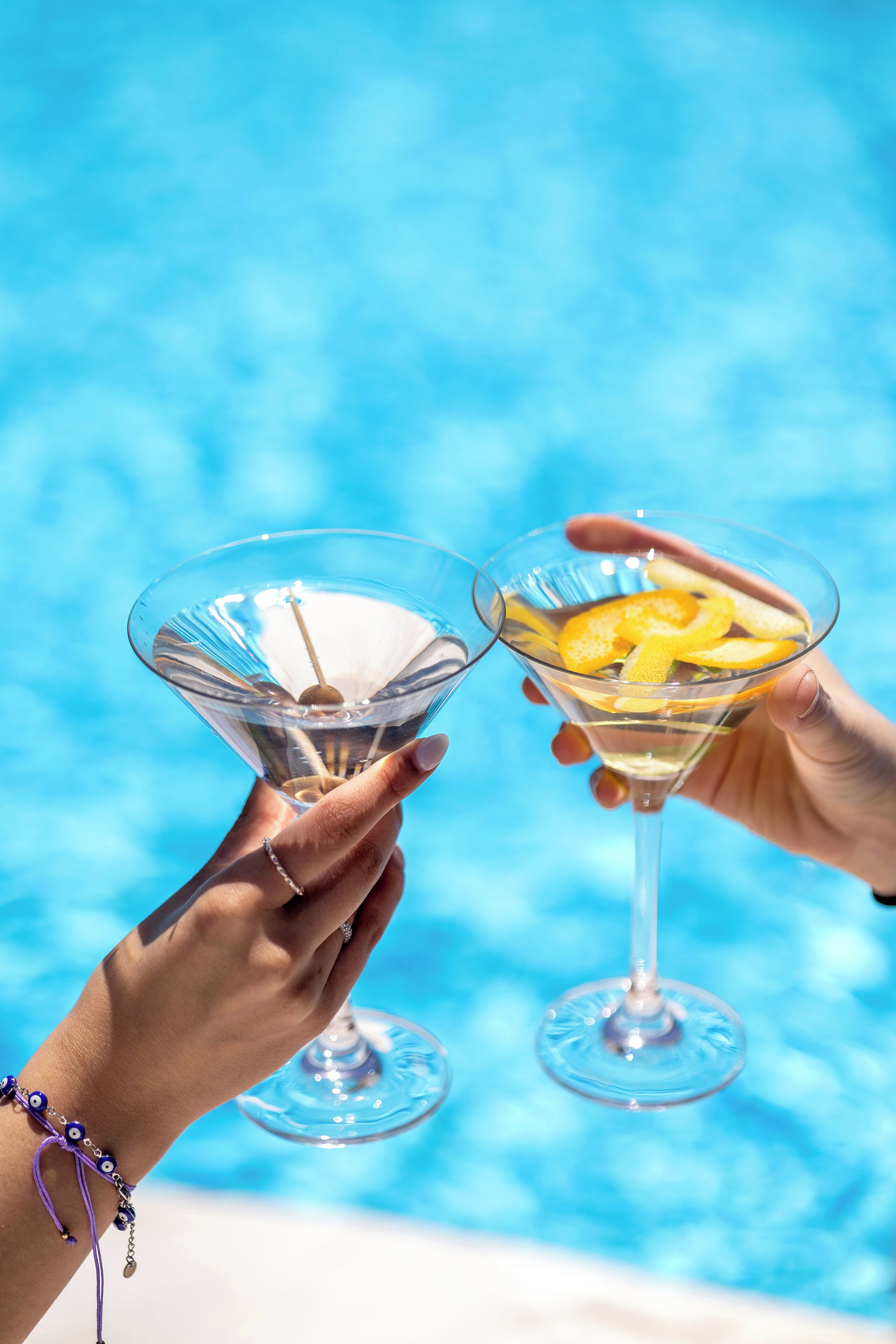 Dos manos sosteniendo copas de martini con una piscina al fondo.