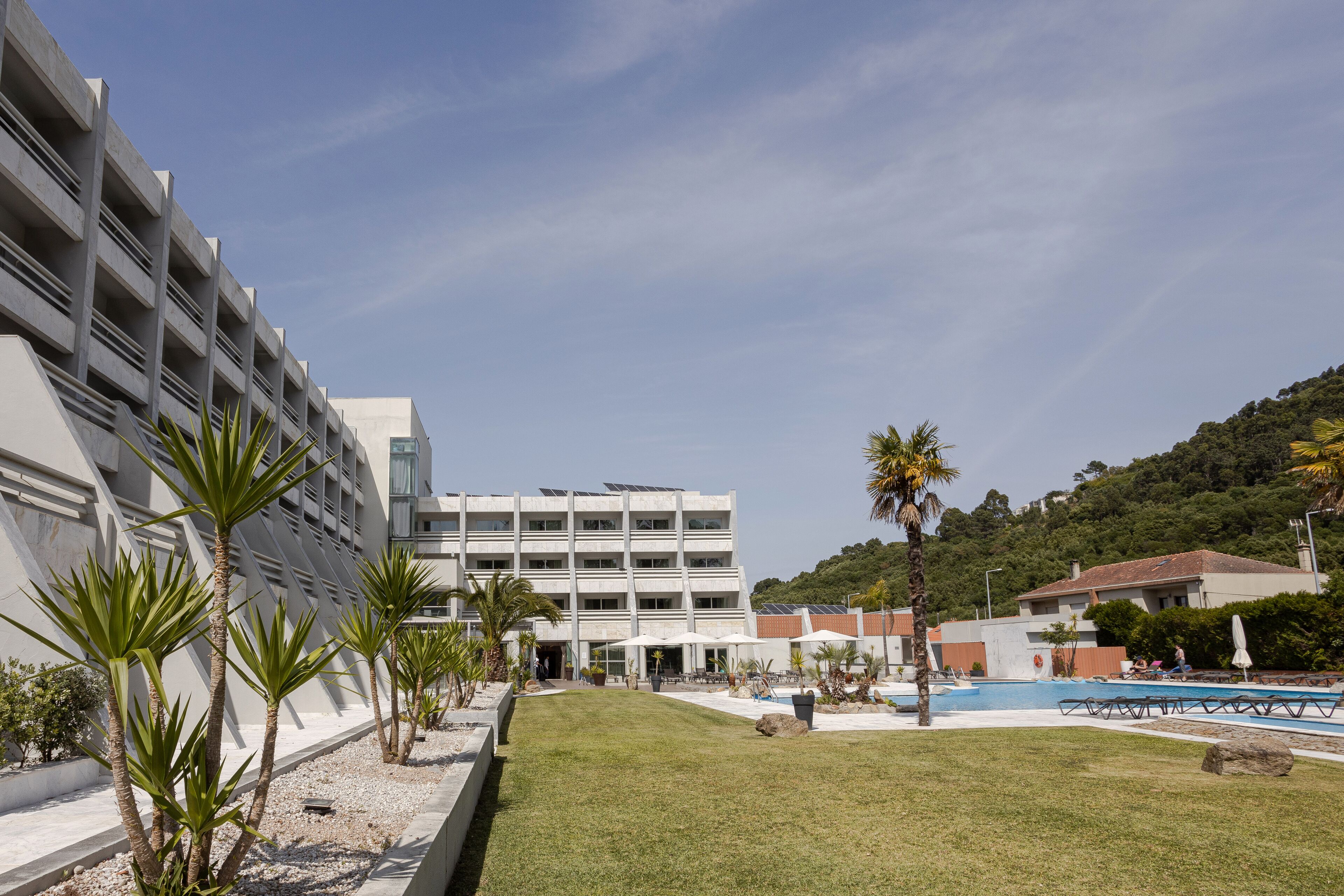 A modern hotel with a pool area, palm trees, and a view of a hilly landscape.