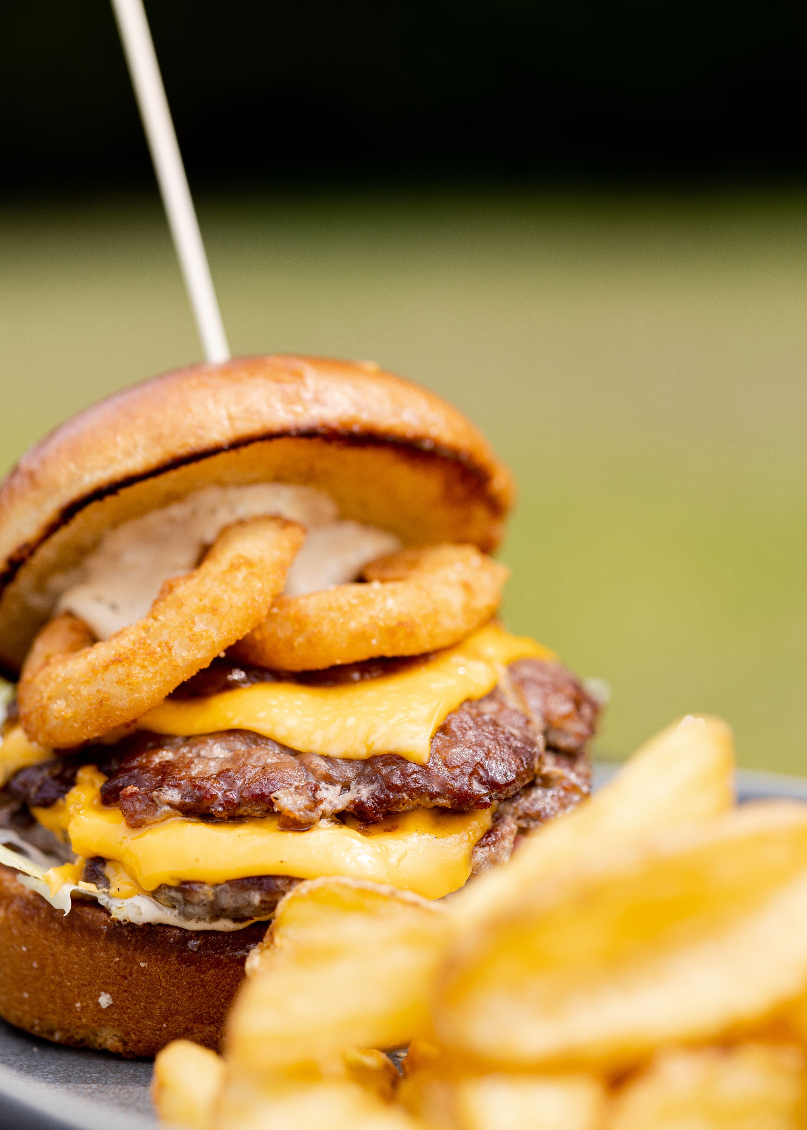 Primer plano de una hamburguesa con queso, carne y aros de cebolla, acompañada de papas fritas.