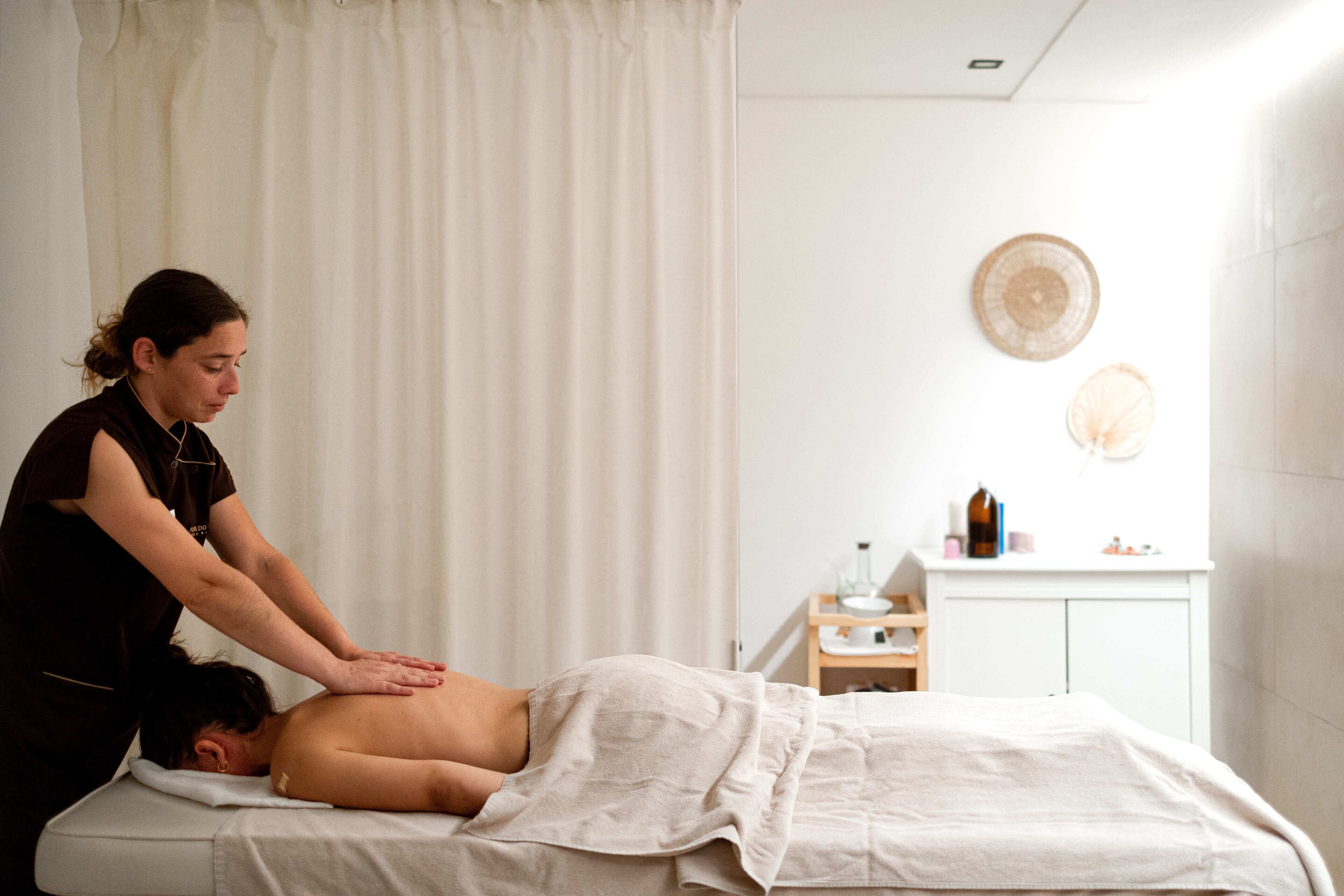 A massage therapy session in a room with a bed, a curtain, a wall-mounted shelf with bottles, and a cabinet.