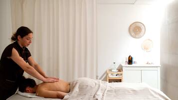 A massage therapy session in a room with a bed, a curtain, a wall-mounted shelf with bottles, and a cabinet.