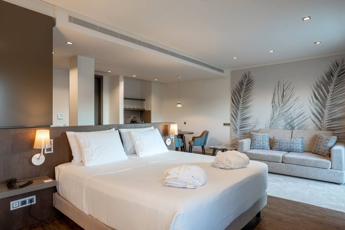 A hotel room with a large bed, a sofa, a dining area, and a bathroom visible through an open door.