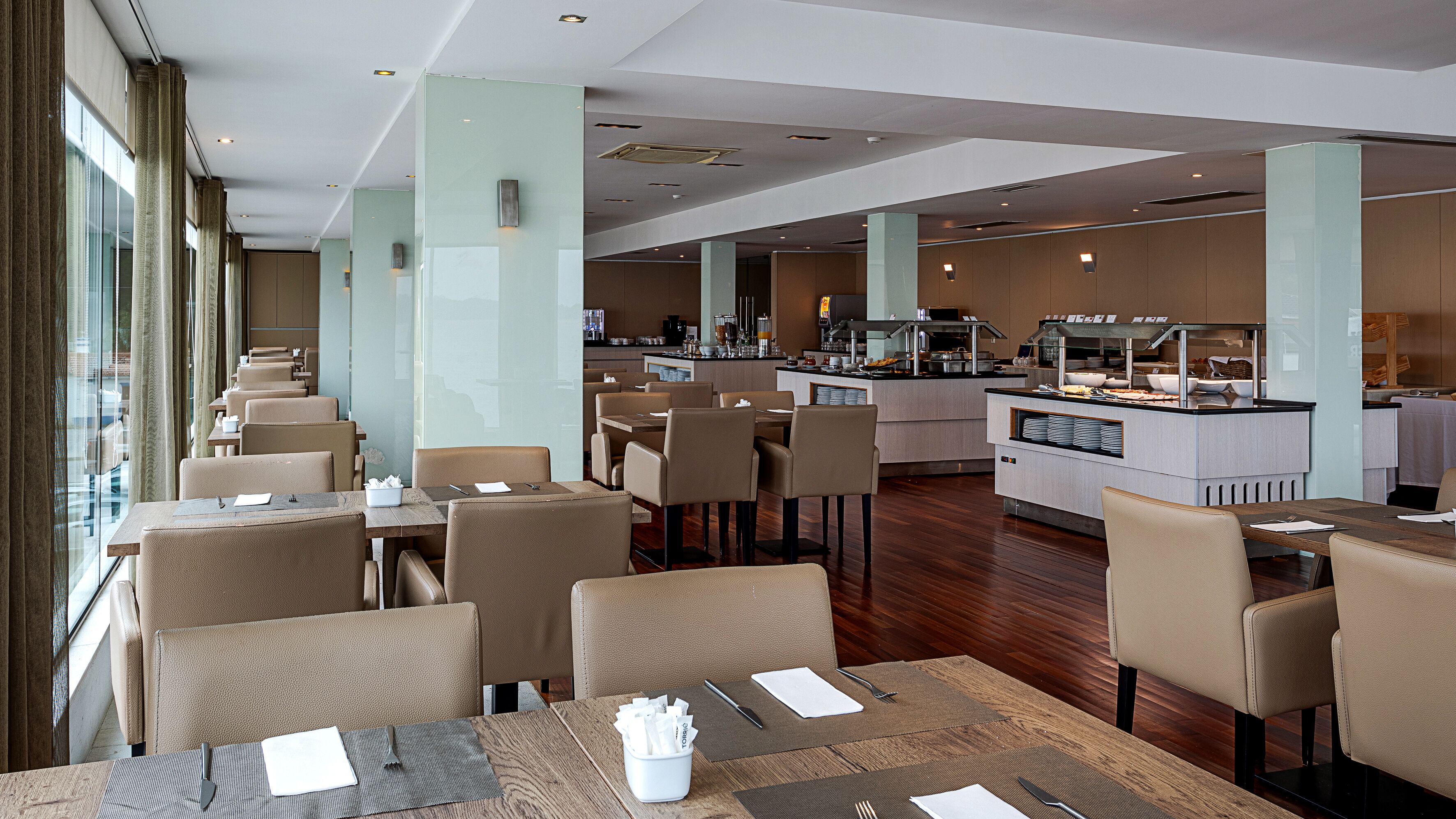 A modern hotel dining area with multiple tables and chairs, a central kitchen, and large windows.