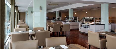 A modern hotel dining area with multiple tables and chairs, a central kitchen, and large windows.