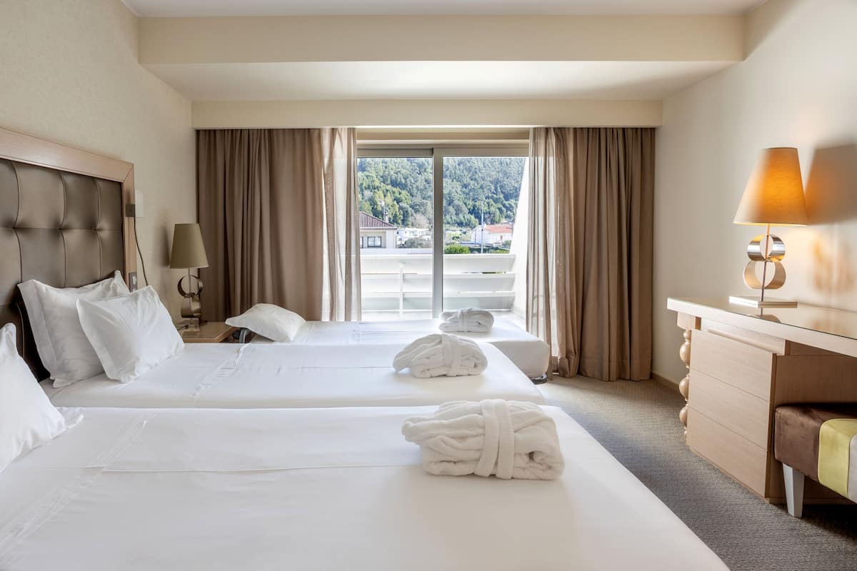 A hotel room with two beds, a desk, a lamp, and a view of a green landscape through the window.