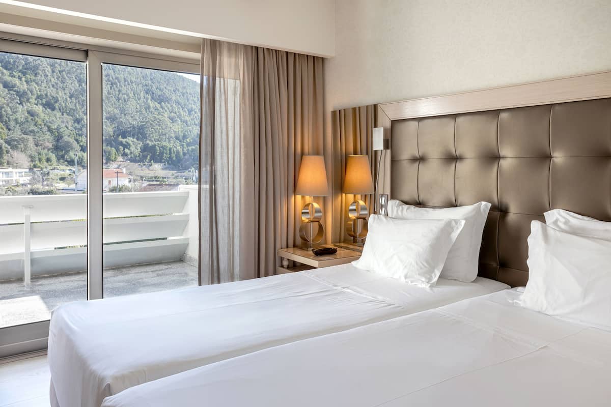 A hotel room with a large bed, two bedside lamps, a nightstand, and a view of a balcony with a mountain backdrop.