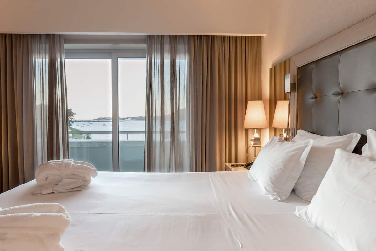A hotel room with a large bed, white linens, a view of the sea, and a balcony.