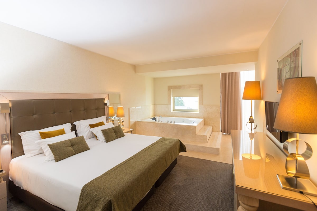 A hotel room with a large bed, a desk, a TV, a bathroom with a bathtub, and a window.